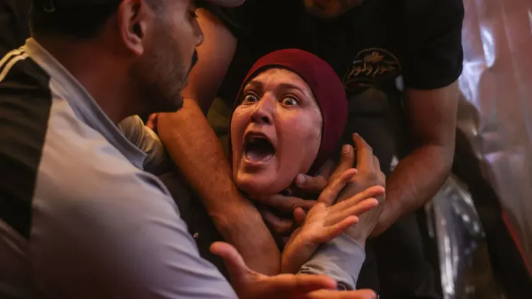 امرأة فلسطينية تعبر عن ألمها الشديد وسط مشهد مأساوي، حيث يحيط بها رجال يحاولون دعمها في ظل التصعيد العسكري.