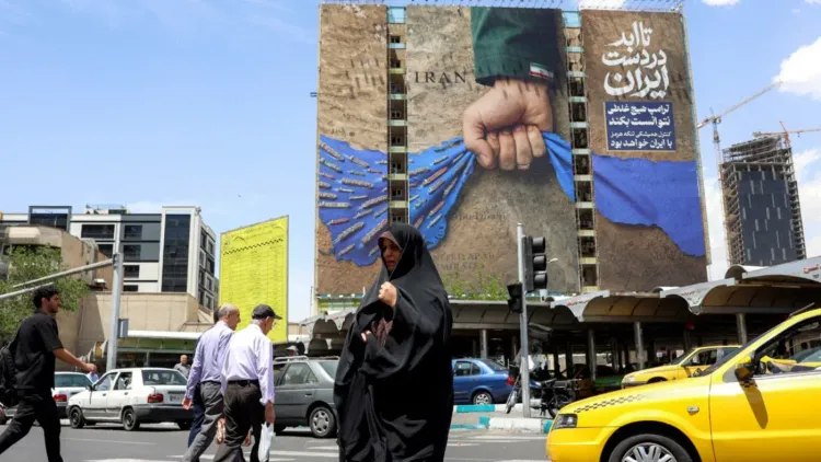 لوحة إعلانات ضخمة في طهران تظهر يدًا ممسكةً بعلم إيران، مع خلفية زرقاء وأسلحة، بينما يمر المارة والسيارات في الشارع.