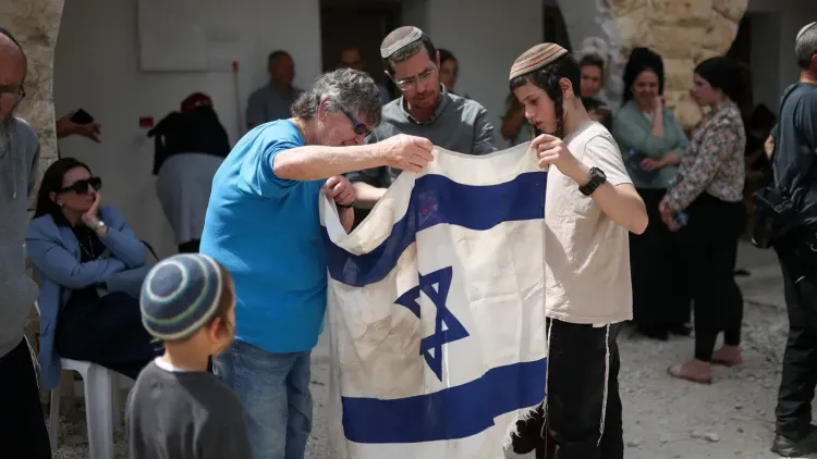 مجموعة من الأشخاص في تجمع، يحملون علمًا إسرائيليًا متسخًا، في سياق مناقشة حول العنف ضد الفلسطينيين في الضفة الغربية.