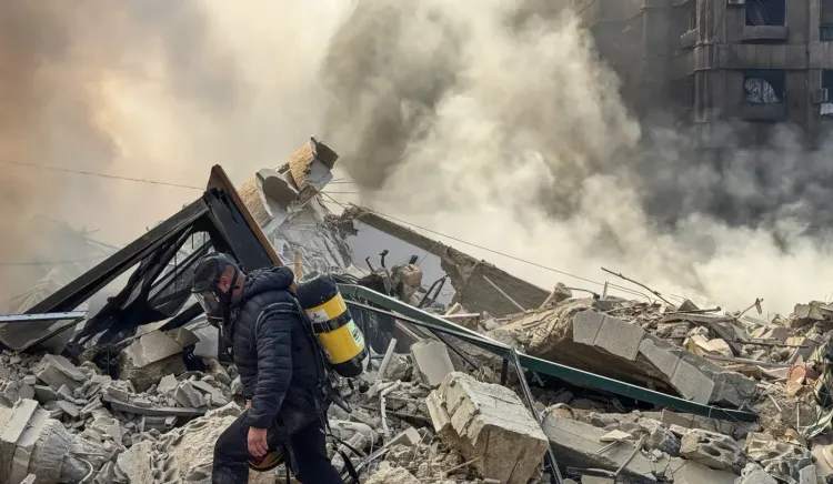 رجل يرتدي سترة واقية يسير بين الأنقاض والدخان بعد هجوم على بيروت، مما يعكس تصاعد التوترات بين حزب الله وإسرائيل.