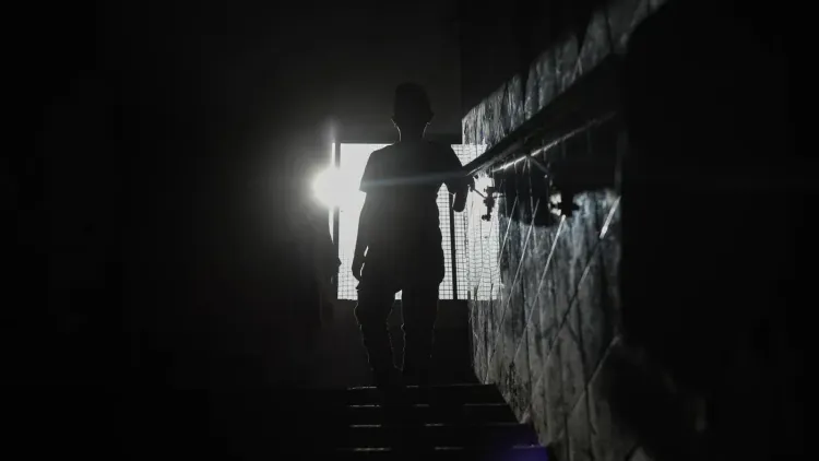 شخص يقف في الظلام أمام نافذة مضاءة، يعكس تحديات انقطاع الكهرباء في غزة وتأثيره على الحياة اليومية.