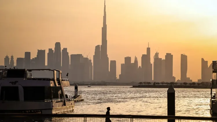 أفق مدينة دبي عند الغروب، يظهر برج خليفة كأعلى نقطة في الأفق، مع قوارب راسية على الواجهة المائية، مما يعكس تأثير الأحداث الأخيرة على الاقتصاد.