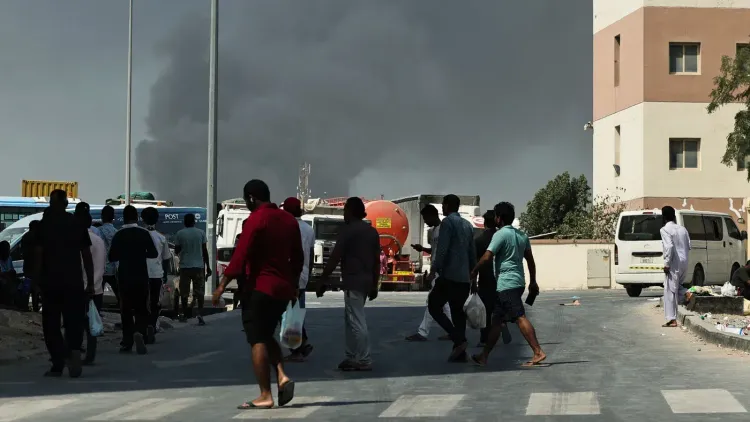 سكان يتجولون في منطقة حضرية، مع تصاعد الدخان في الخلفية نتيجة الغارات الجوية، مما يعكس الوضع المتأزم في الخليج.