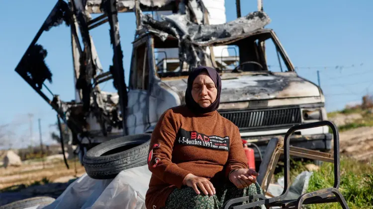 امرأة جالسة أمام شاحنة محترقة في منطقة متضررة، تعكس آثار الصراع في الأراضي الفلسطينية المحتلة وتسلط الضوء على معاناة المدنيين.