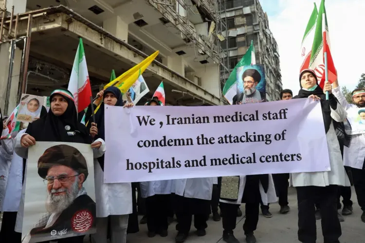موظفو القطاع الطبي الإيراني يحتجون ضد الهجمات على المستشفيات والمراكز الطبية، حاملين لافتة تدين الاعتداءات، مع خلفية لمباني متضررة.