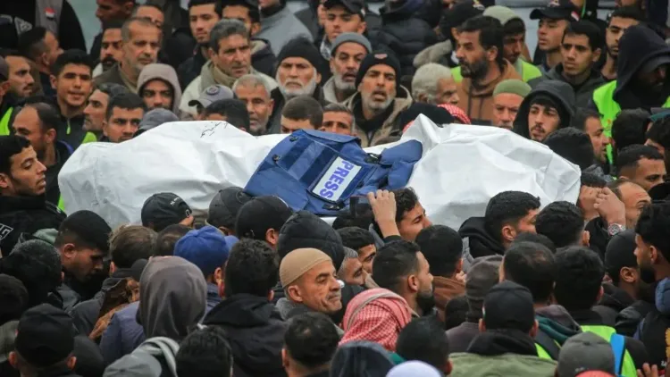 تجمع حشد كبير من الناس حول جثمان صحفي مغطى بكيس أبيض يحمل علامة "صحافة"، مما يعكس تأثير العنف على الإعلاميين الفلسطينيين.
