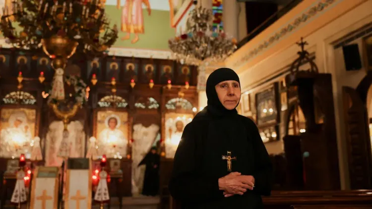 امرأة ترتدي زيًا دينيًا تقف في كنيسة، محاطة بأيقونات مضاءة وشمعدانات، تعكس التوترات المستمرة في المجتمع المسيحي في المنطقة.