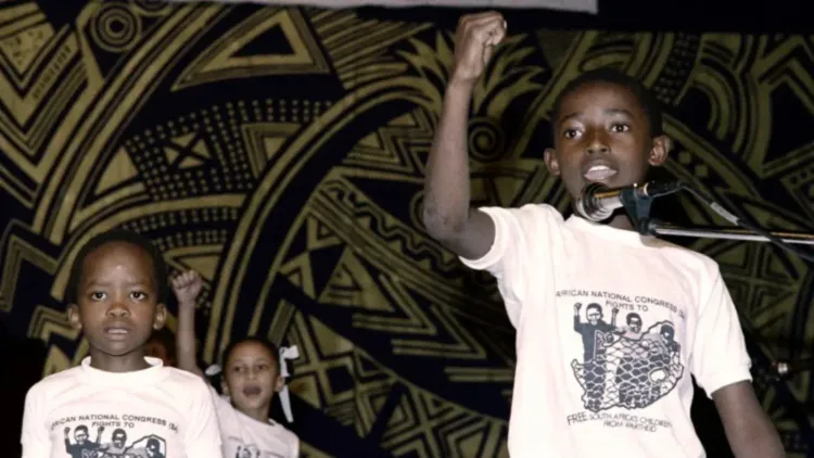 أطفال يتحدثون في حدث عام، يرتدون قمصان تحمل شعار "المؤتمر الوطني الأفريقي"، يعبرون عن مقاومة الظلم والمعاناة.