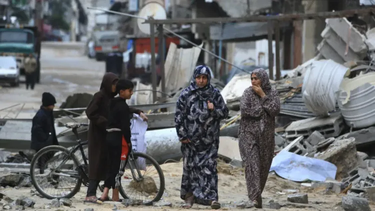 نساء وأطفال يسيرون في منطقة مدمرة في غزة، مع بقايا المنازل المهدمة حولهم، مما يعكس آثار الصراع المستمر.