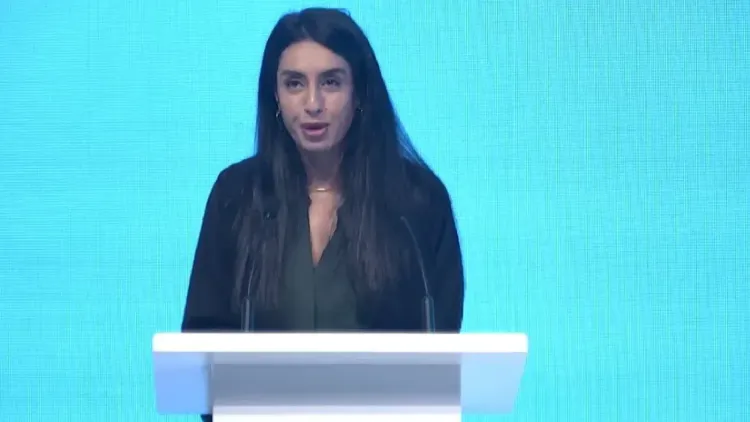 هند العويس تتحدث على منصة في حدث رسمي، مع خلفية زرقاء، حيث تدافع عن نفسها في سياق الجدل حول رسائلها مع جيفري إبشتاين.