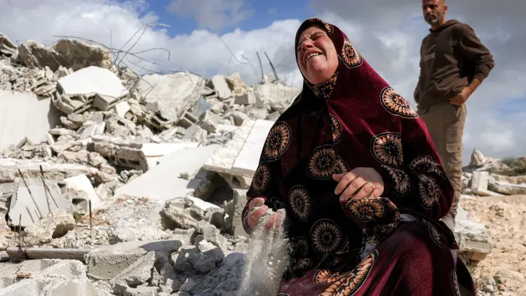 امرأة فلسطينية تجلس على أنقاض منزلها المهدوم، تعبر عن حزنها الشديد بينما يراقبها رجل خلفها، في سياق التهجير القسري في الضفة الغربية.