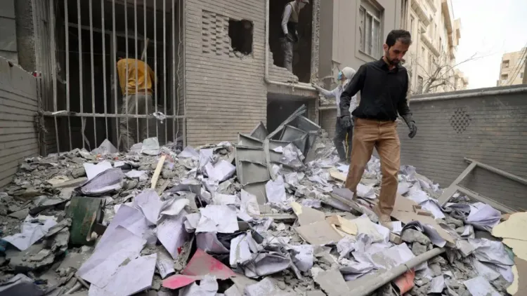 رجل يمشي بين أنقاض مبنى مدمر، محاط بأوراق ممزقة وحطام، يعكس آثار النزاع في المنطقة.