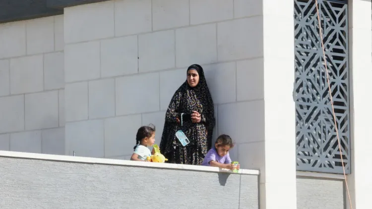 امرأة فلسطينية ترتدي عباءة تقف على شرفة منزل محاصر في قصرة مع طفلين، تعكس معاناة الحصار ونقص الغذاء في الضفة الغربية.