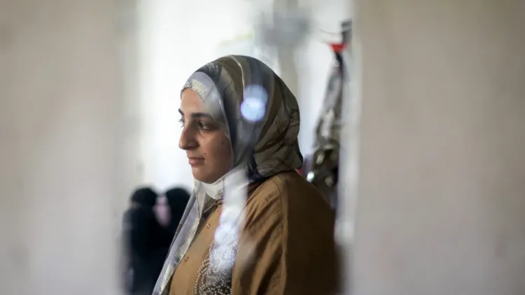 شابة فلسطينية ترتدي الحجاب تقف بجانب نافذة في غزة، تعبيرها يعكس الصبر والأمل وسط ظروف صعبة للتعليم والحياة.