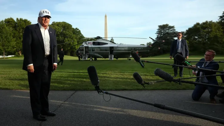 ترامب يرتدي قبعة مكتوب عليها "USA" ويتحدث إلى الصحفيين في حديقة البيت الأبيض، مع وجود مروحية خلفه ونصب واشنطن في الخلفية.