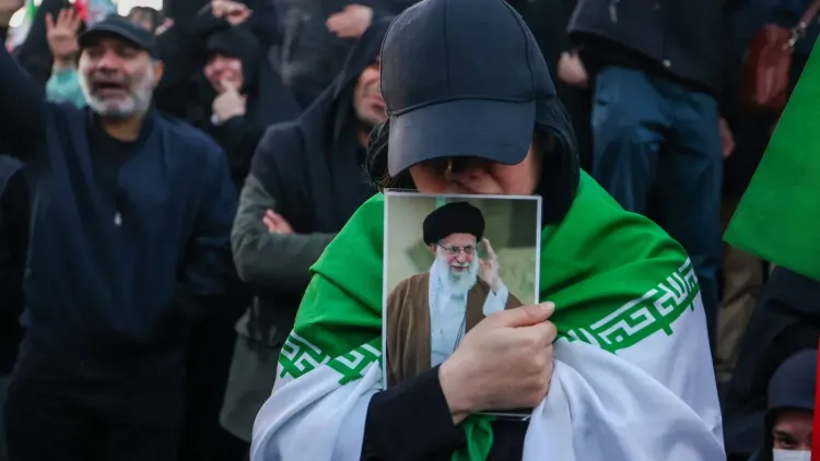 امرأة تحمل صورة للمرشد الأعلى الإيراني علي خامنئي، ترتدي علمًا إيرانيًا، وسط حشد من المتظاهرين. تعكس الصورة مشاعر التأييد والاحتجاج في سياق التوترات السياسية.