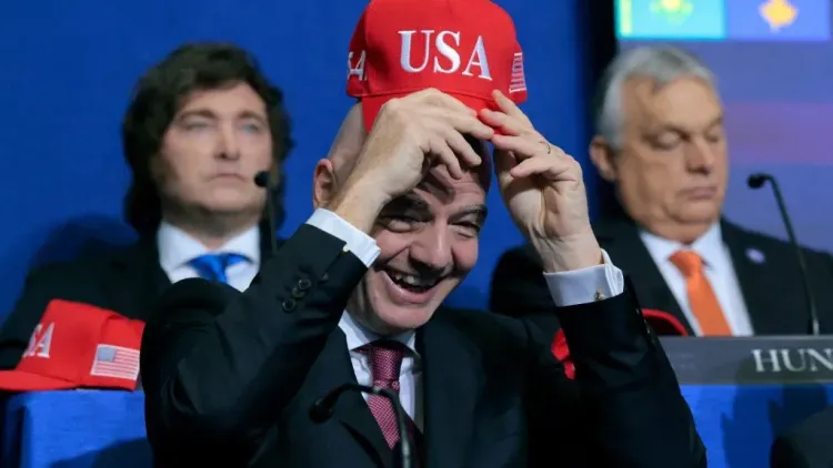 رئيس FIFA جاني إنفانتينو يبتسم وهو يرتدي قبعة مكتوب عليها USA خلال مؤتمر صحفي، مع وجود شخصين خلفه في الخلفية.