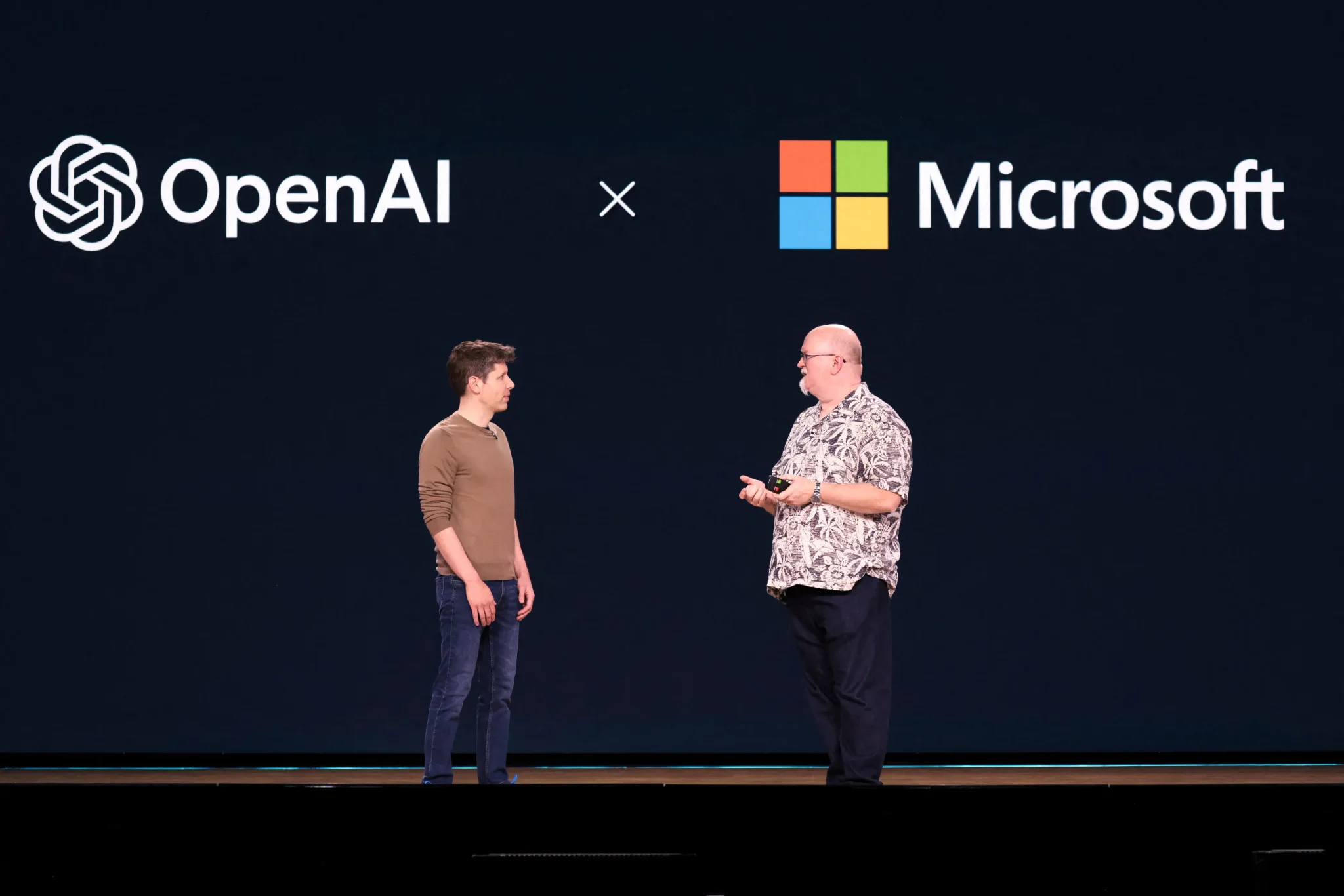 شخصان يتحدثان أمام خلفية شعار OpenAI وشعار Microsoft، تعبير عن الشراكة والتنافس في مجال الذكاء الاصطناعي والإيرادات المشتركة.