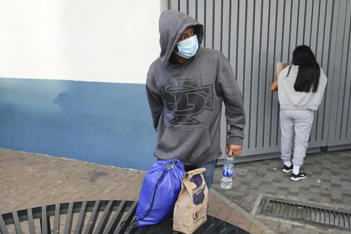 شخص يرتدي قناع وجه ويقف بجانب طاولة حديدية في منطقة حضرية، يحمل حقيبة زرقاء وزجاجة مياه، بينما تظهر امرأة خلفه.
