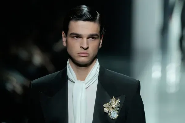 عارض أزياء يرتدي بدلة أنيقة مع ربطة عنق بيضاء، ويزين صدره بمجوهرات فاخرة، يعكس الاتجاهات الحديثة في الموضة الرجالية.