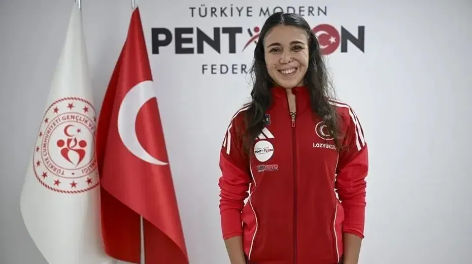 الرياضية التركية İlke Özyüksel Mihrioğlu مبتسمة ترتدي زيًا رياضيًا أحمر أمام علمي تركيا وشعار الاتحاد التركي للخماسي الحديث.