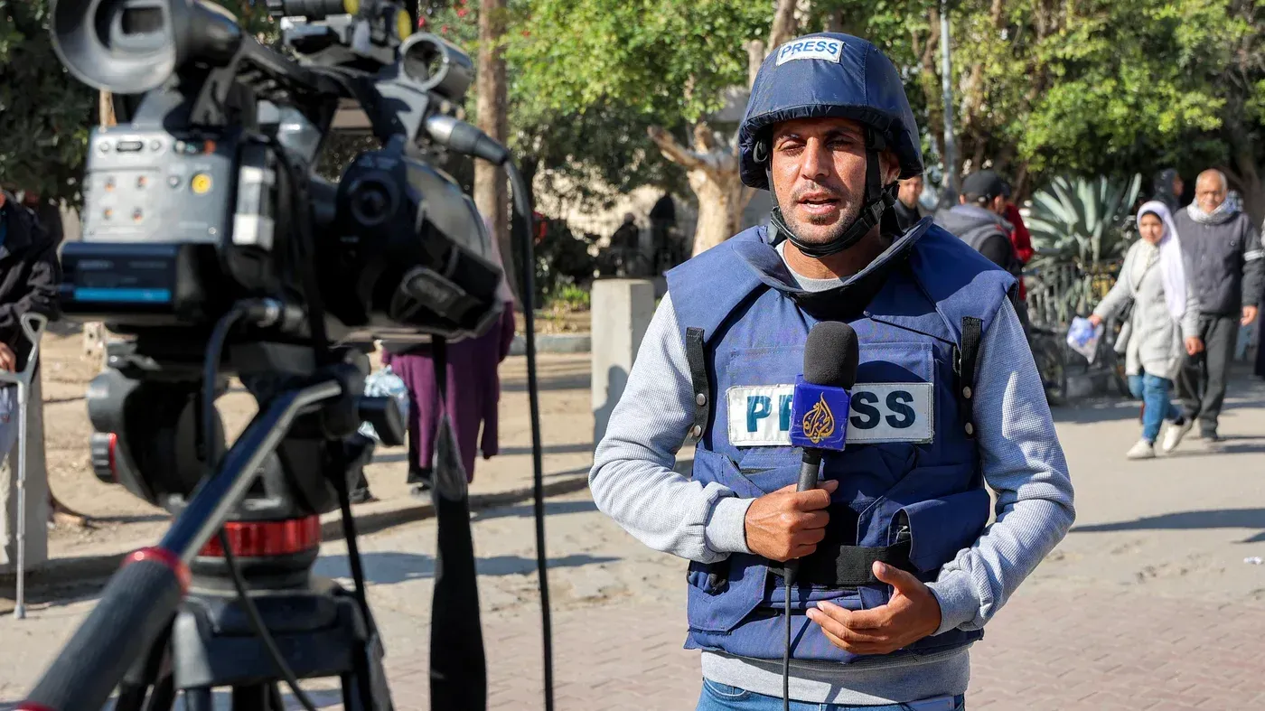 محمد قريقع، مراسل قناة الجزيرة، يتحدث أمام الكاميرا مرتديًا سترة صحفية، مع خلفية تشير إلى الأوضاع في غزة.