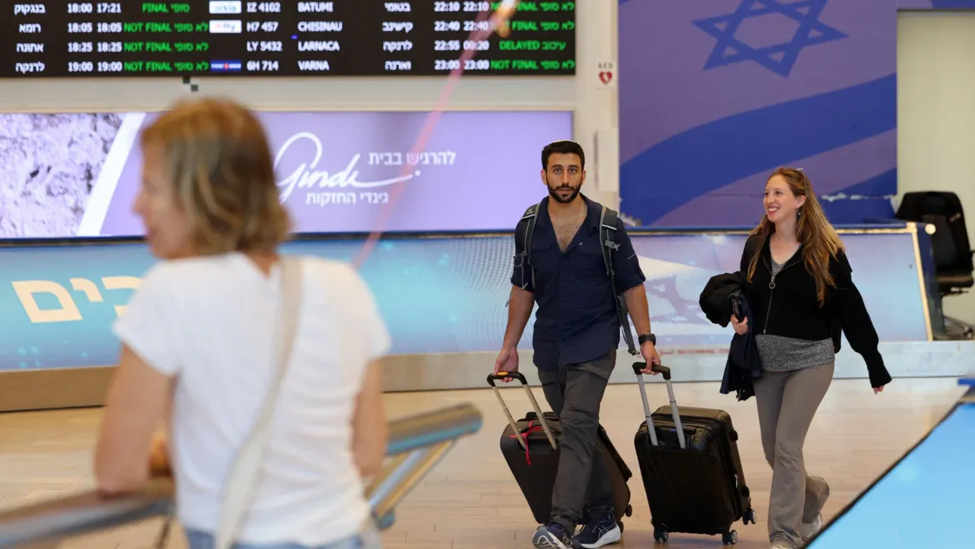 شخصان يحملان حقائب سفر في مطار إسرائيلي، مع شاشة تعرض مواعيد الرحلات في الخلفية، مما يعكس حركة الهجرة من البلاد.