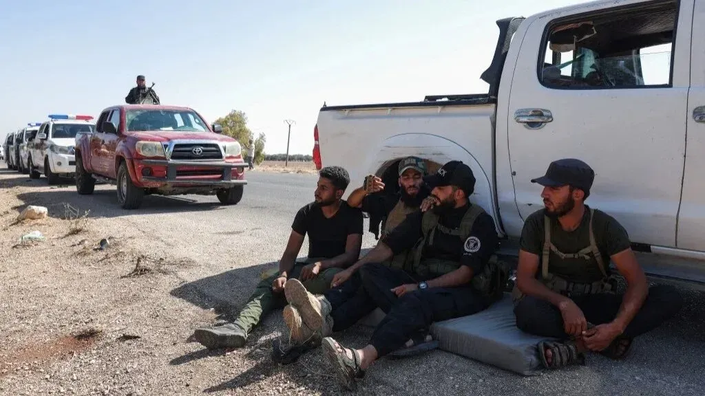 مجموعة من المقاتلين يجلسون على جانب الطريق في السويداء، مع وجود سيارات عسكرية في الخلفية، وسط أجواء من التوتر بعد القتال العنيف.