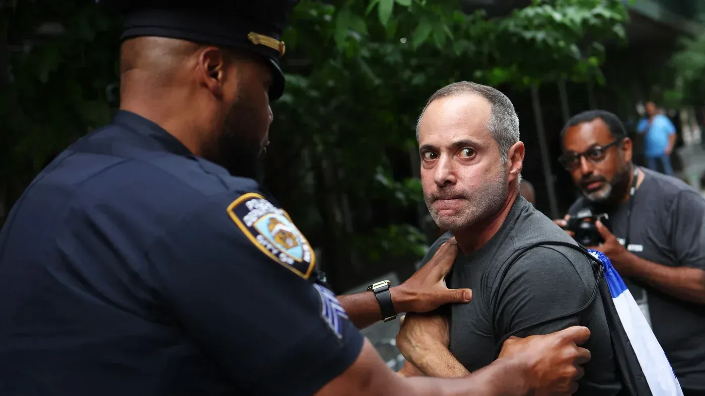 شخص يتعرض للاحتجاز من قبل شرطي في نيويورك، وسط مظاهرة تتعلق بقضايا كراهية. تعكس الصورة التوترات الاجتماعية حول العدالة.