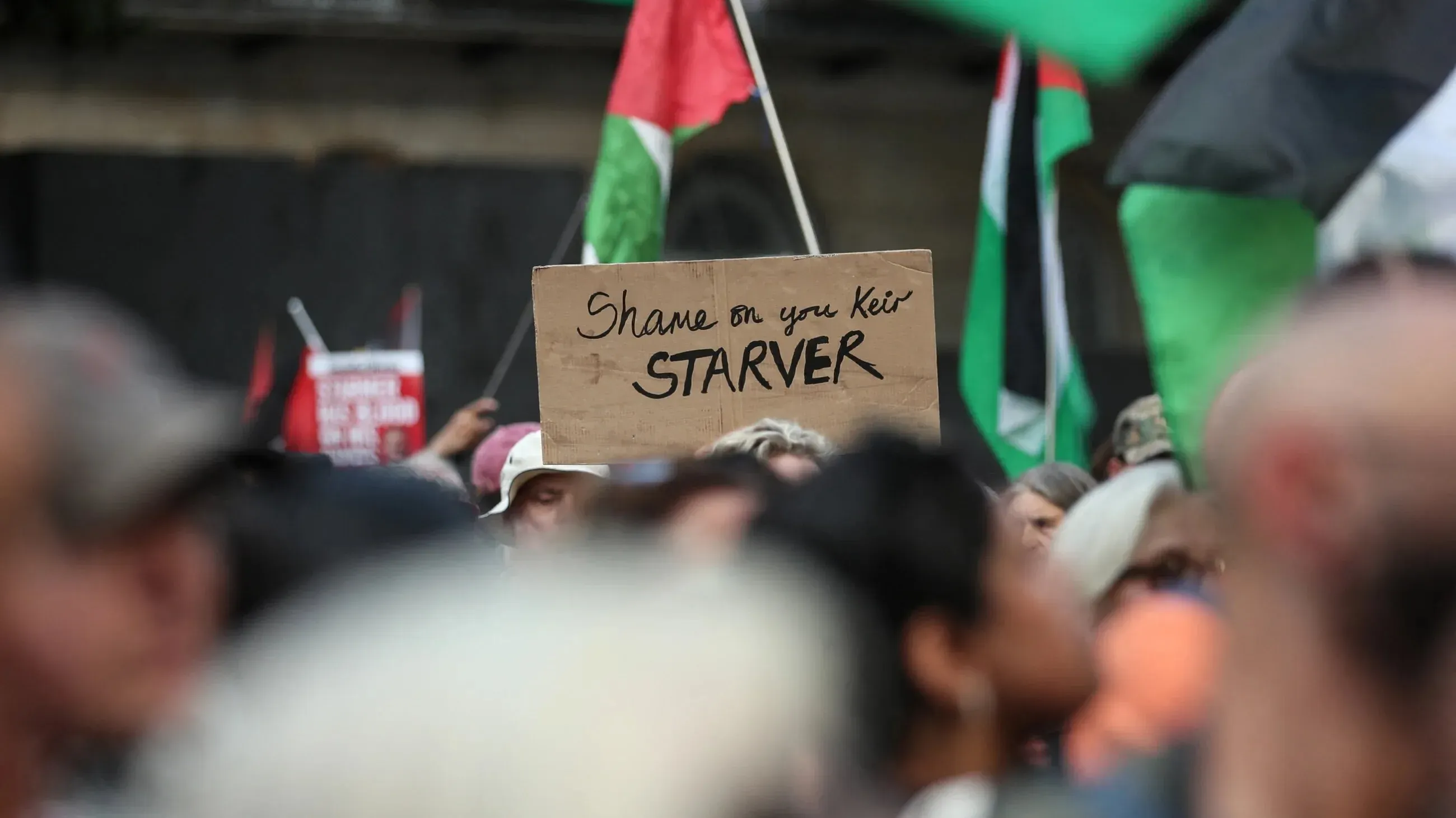 لافتة في مظاهرة تشير إلى استياء المتظاهرين من كير ستارمر، محاطة بأعلام فلسطينية. تعكس الشعارات المطالبة بالاعتراف بالدولة الفلسطينية.