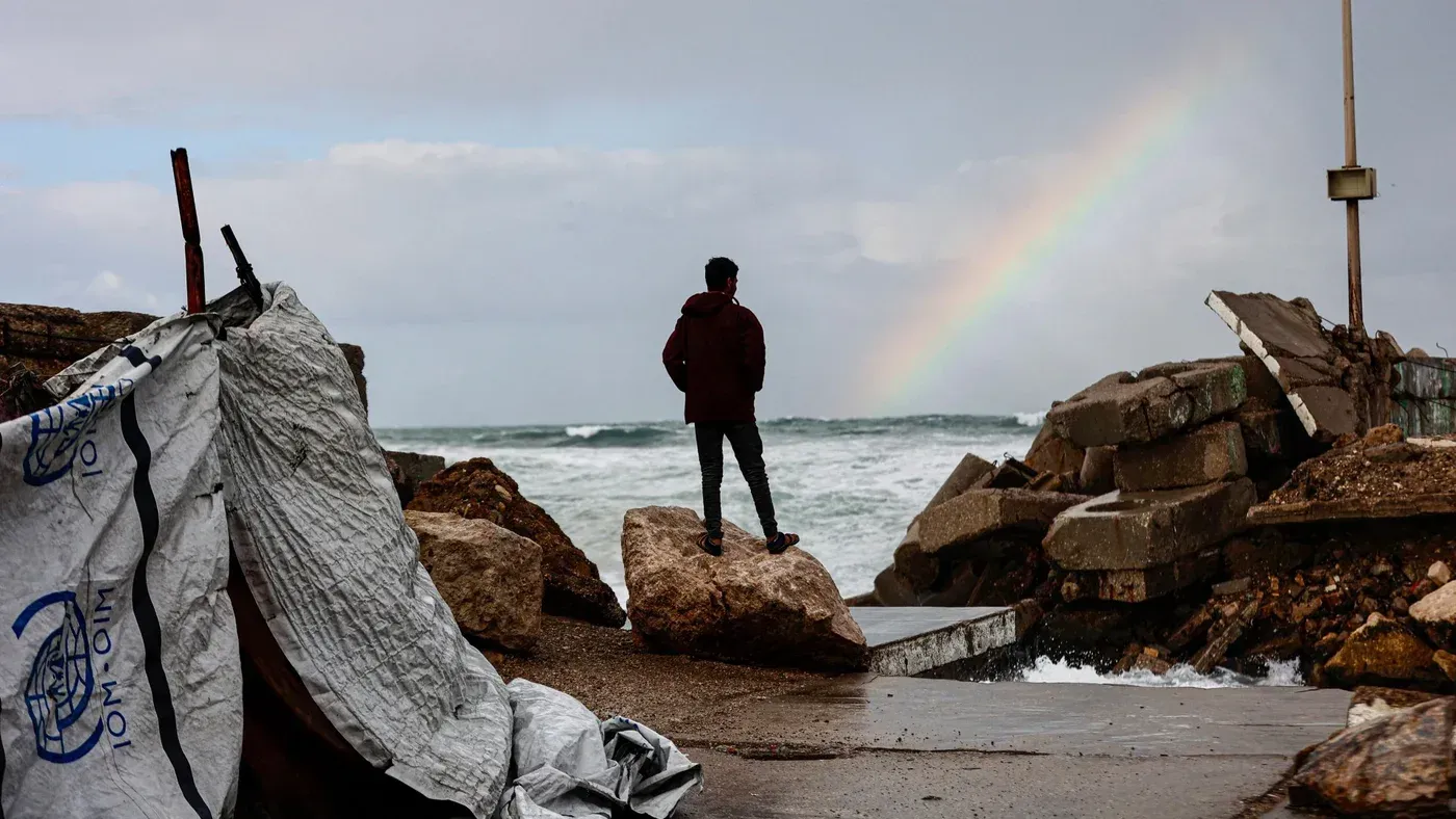 شاب يقف على حافة البحر في غزة، يراقب الأمواج تحت قوس قزح، بينما تظهر آثار الدمار من العواصف الأخيرة.