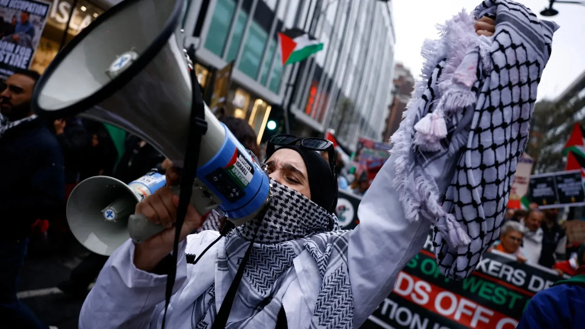 امرأة ترتدي الكوفية وتحمل مكبر صوت خلال مظاهرة لدعم فلسطين، محاطة بأعلام فلسطينية ولافتات تطالب بالعدالة.