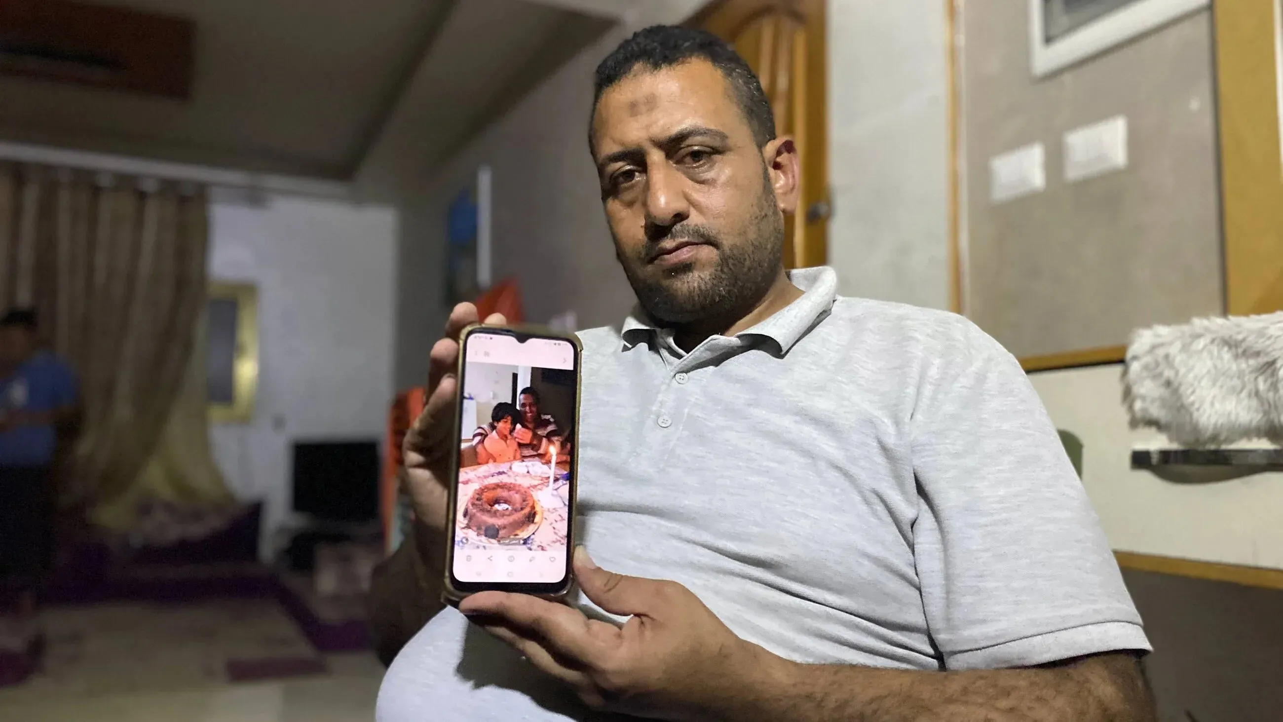 والد نوح السقا يحمل هاتفه الذكي الذي يظهر صورة لابنه نوح يحتفل بعيد ميلاده مع كعكة بسيطة، وسط أجواء من الحزن بسبب الظروف الصعبة في غزة.