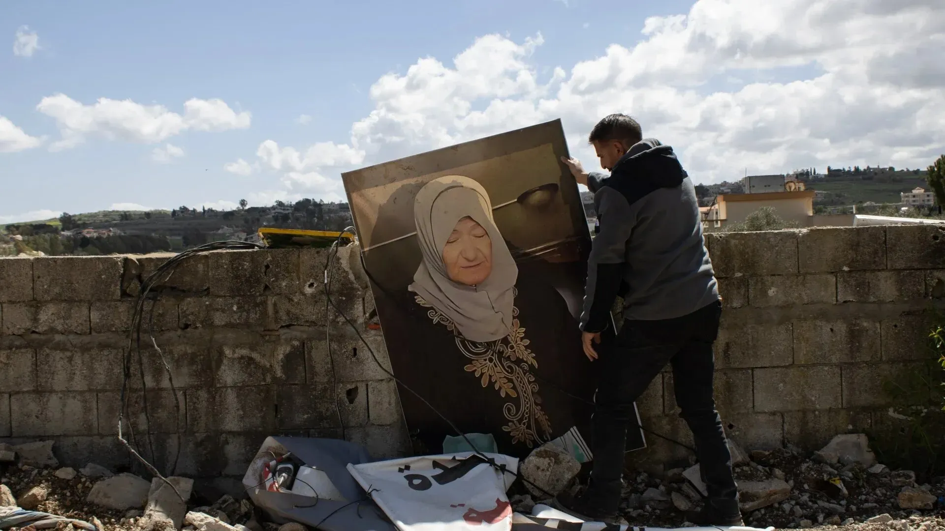 ابن يحمل صورة كبيرة لوالدته الراحلة نجوى غشام، بينما يقف أمام أنقاض منزلها في قرية يارون، جنوب لبنان.