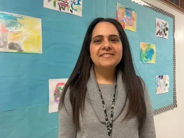 مساعدة تعليمية مبتسمة تقف أمام جدار مزين برسومات أطفال، تعكس جهود تحسين التعليم في مرحلة الطفولة المبكرة في ميشيغان.