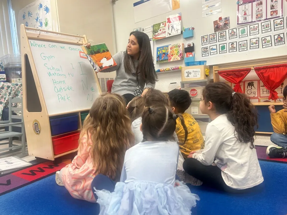 معلمة تشرح درسًا للأطفال في فصل دراسي لرياض الأطفال، حيث يجلس الأطفال في دائرة ويستمعون بانتباه.