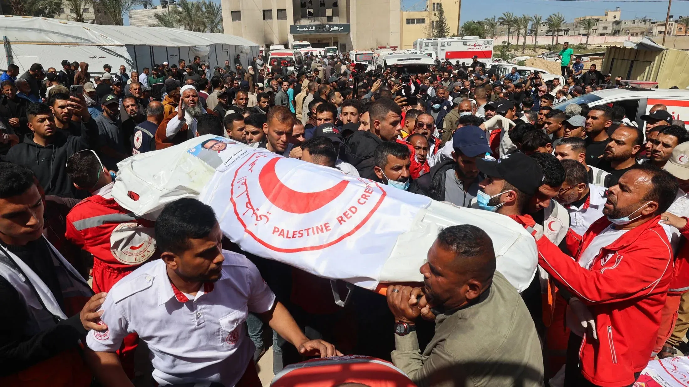 مئات من الناس يشاركون في جنازة عمال الإنقاذ الفلسطينيين، حاملين نعشًا مغطى بشعار الهلال الأحمر، في مشهد يعكس الحزن والغضب.