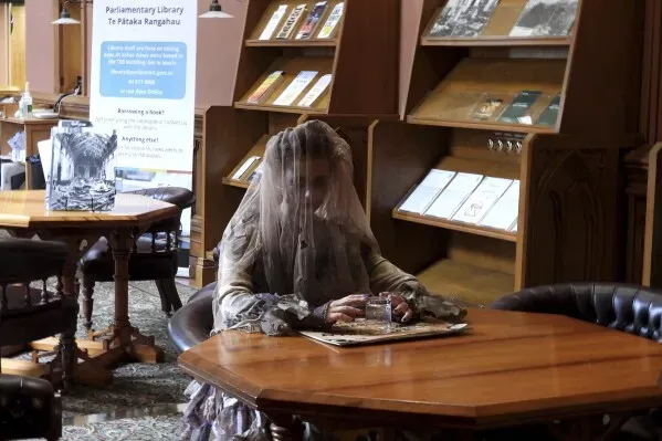 امرأة ترتدي زيًا فيكتوريًا مع غطاء رأس شفاف تجلس على طاولة في مكتبة البرلمان، محاطة بكتب وأثاث قديم، تعكس أجواءً تاريخية وغامضة.