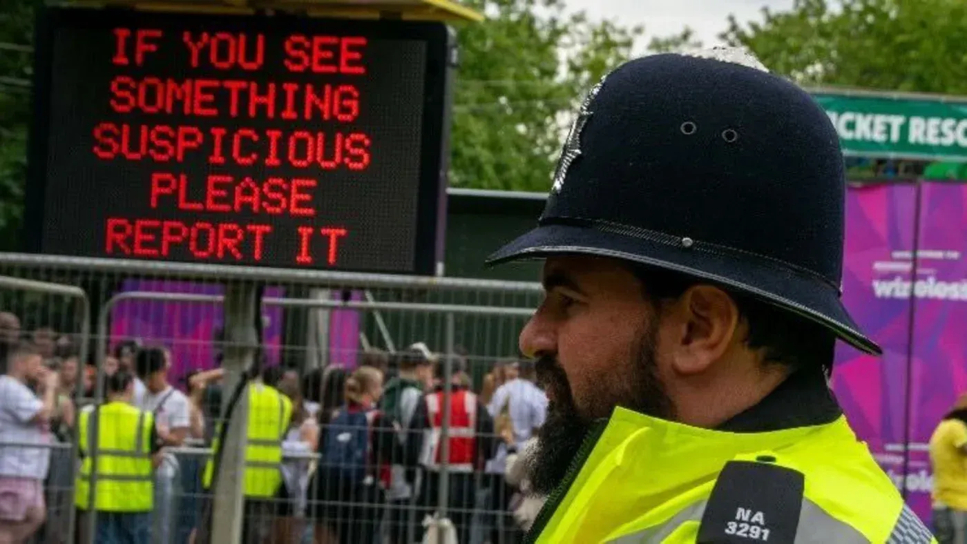 شرطي يرتدي زيًا رسميًا يقف بجوار لافتة إلكترونية تطلب من الناس الإبلاغ عن أي شيء مشبوه، مما يعكس جهود مكافحة الإرهاب.