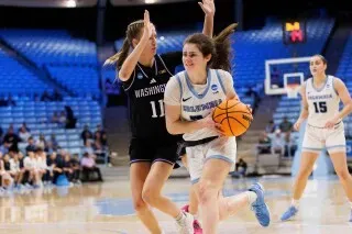 لاعبة كرة سلة تتقدم بالكرة خلال مباراة، بينما تحاول لاعبة من فريق واشنطن الدفاع عنها. المقاعد في الخلفية فارغة.