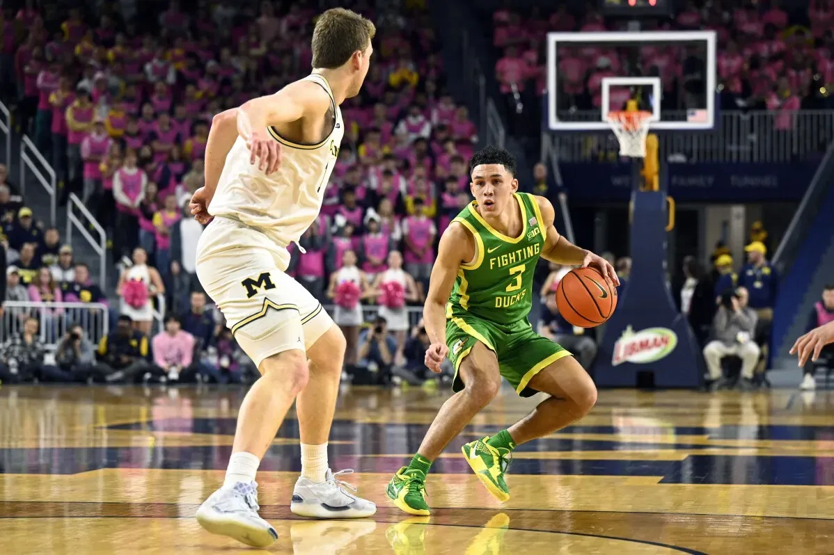 لاعب كرة سلة من فريق أوريغون يت dribble الكرة بينما يتجه نحو سلة فريق ميشيغان، مع جمهور مكون من مشجعين يرتدون قمصان وردية في الخلفية.