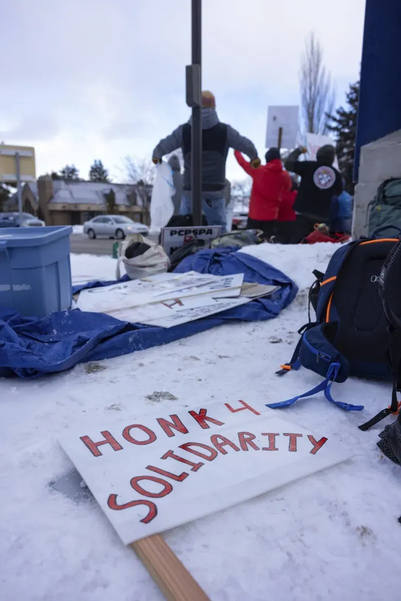 لافتة مكتوب عليها \"HONK 4 SOLIDARITY\" موضوعة على الثلج، مع متظاهرين في الخلفية خلال إضراب عمال دوريات التزلج في بارك سيتي.