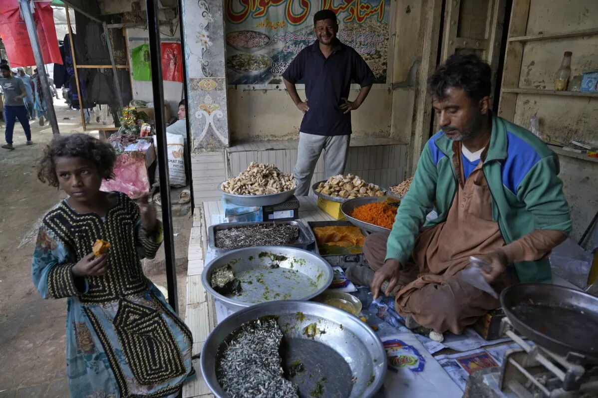 طفلة تأكل ثمرة في سوق جوادر، حيث يقوم بائع بتحضير الطعام، مع وجود رجل آخر خلفه. السوق يعكس الحياة اليومية في المدينة.