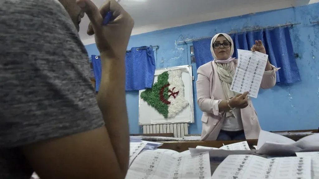 امرأة ترتدي حجابًا، تحمل ورقة اقتراع في مركز انتخابي جزائري، مع خلفية تحتوي على خريطة الجزائر. تعكس الصورة أجواء الانتخابات المقبلة.