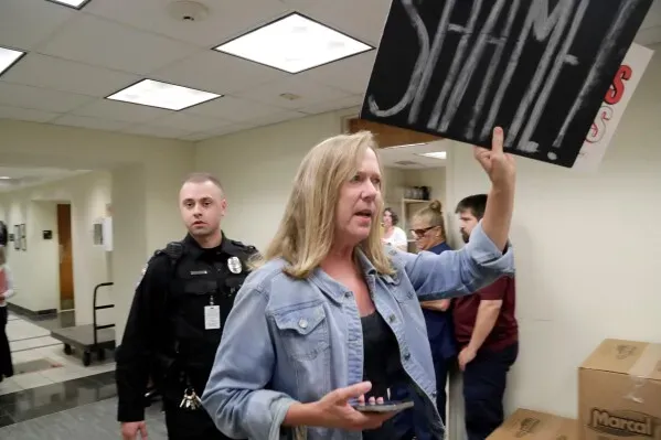 امرأة تحمل لافتة مكتوب عليها "عار" بينما تتحدث، مع وجود ضابط شرطة في الخلفية، في سياق احتجاج حول إعادة رسم دوائر الانتخابات في نورث كارولينا.