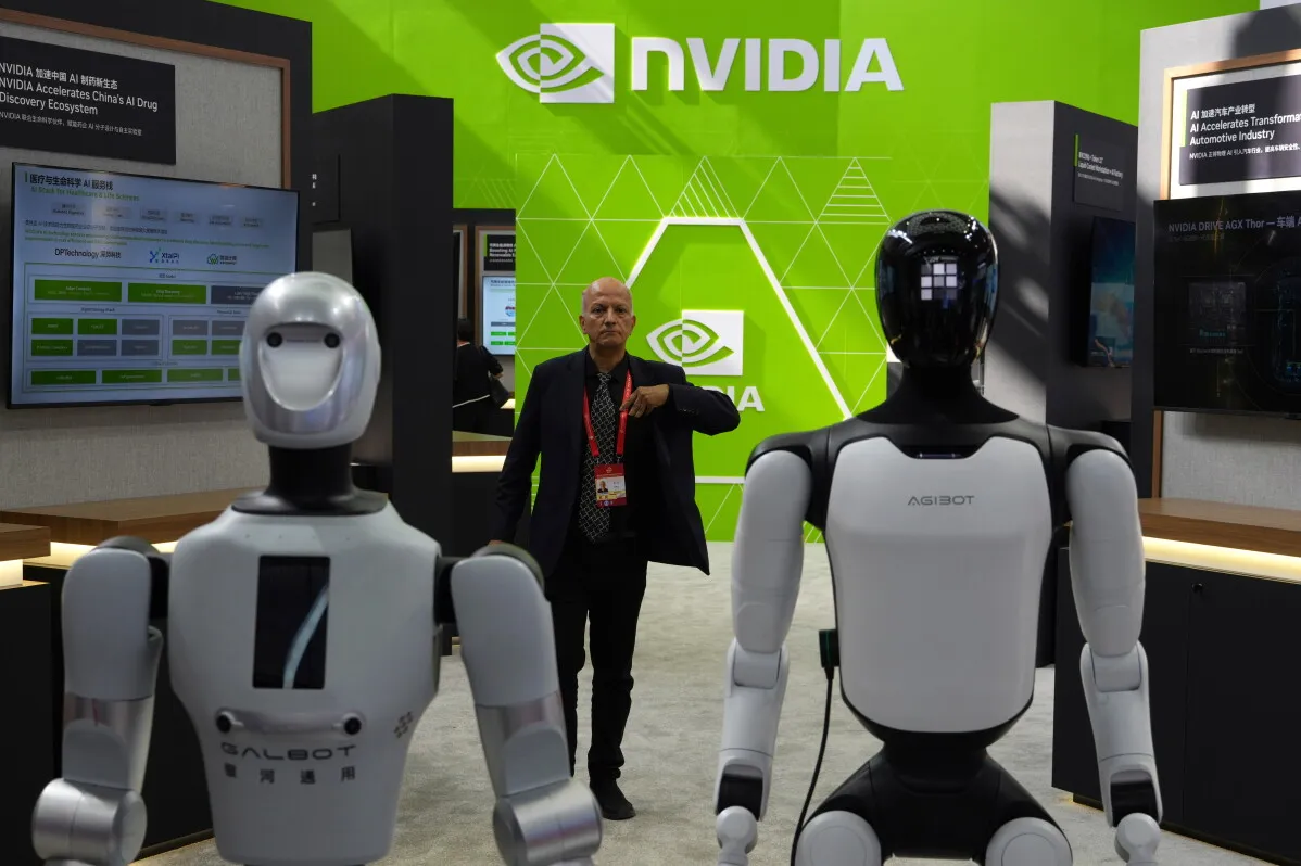 رجل يقف بين روبوتين في معرض لشركة Nvidia، مع شعار الشركة باللون الأخضر في الخلفية، يعكس تنافس التكنولوجيا والذكاء الاصطناعي.