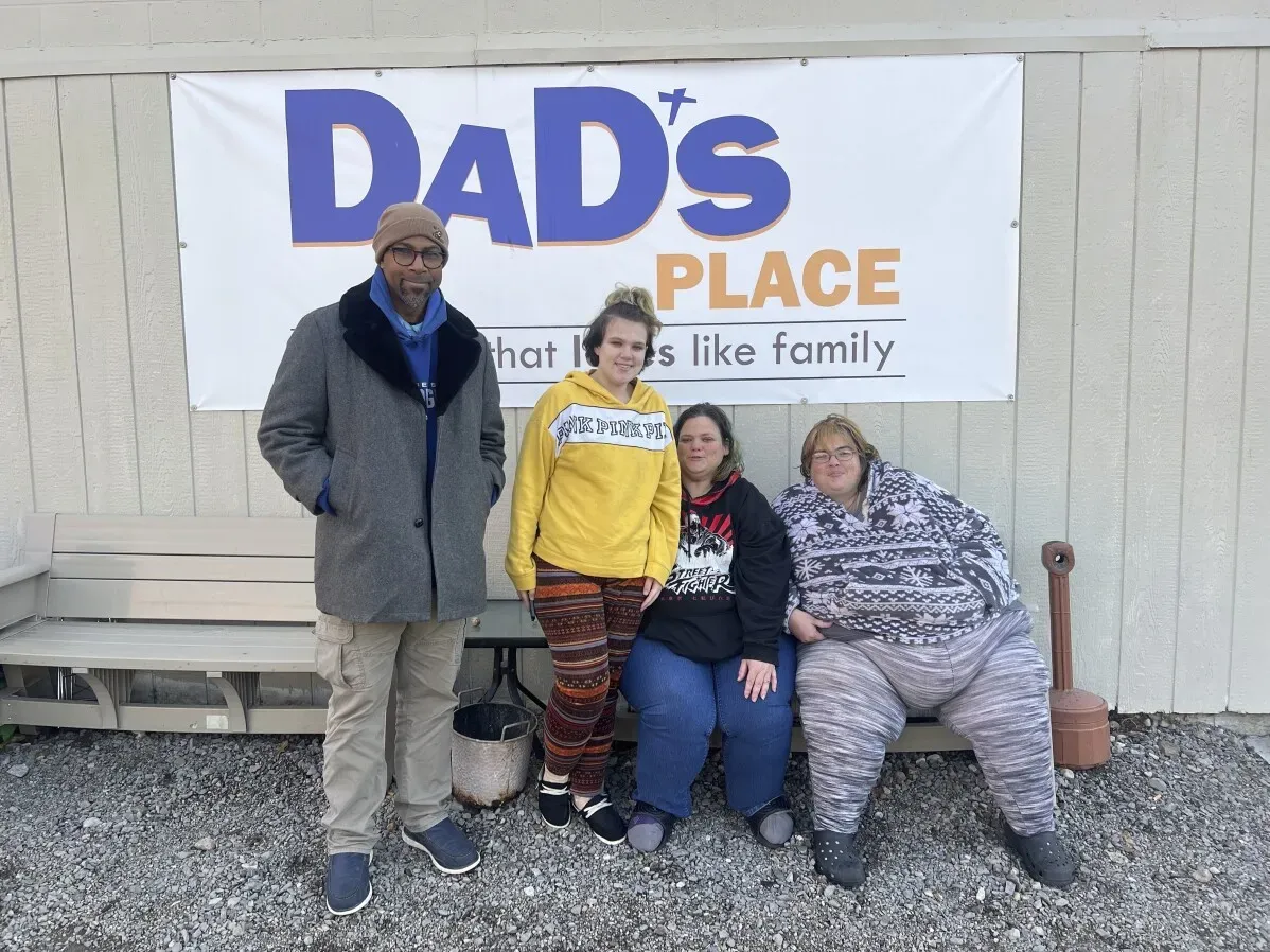 مجموعة من الأشخاص يجلسون على مقعد أمام لافتة تشير إلى "Dad's Place"، حيث يتم توفير مأوى للمشردين في بريان، أوهايو.