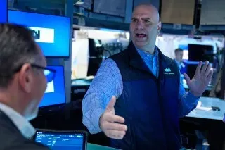 متداول في بورصة نيويورك يتحدث بحماس مع زميله، مع شاشات تعرض بيانات السوق في الخلفية، وسط تقلبات أسعار النفط والأسواق المالية.