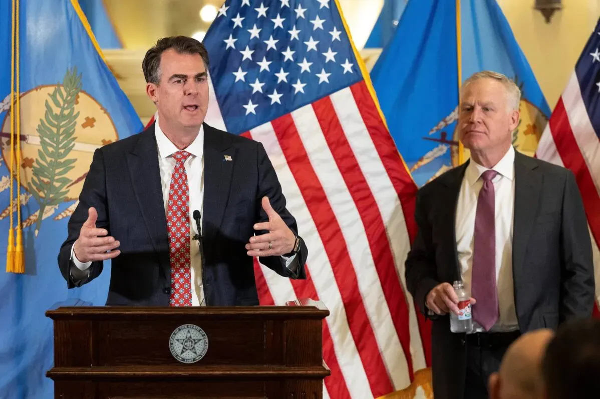 حاكم ولاية أوكلاهوما كيفن ستيت يتحدث خلال مؤتمر صحفي، بينما يقف آلان أرمسترونغ خلفه، مع العلم الأمريكي وعلم الولاية في الخلفية.