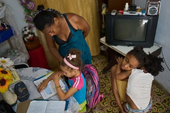 امرأة وطفلتان في غرفة معيشة بسيطة في هافانا، حيث يبدو القلق على وجه الأم بينما تساعد ابنتها في الدراسة.